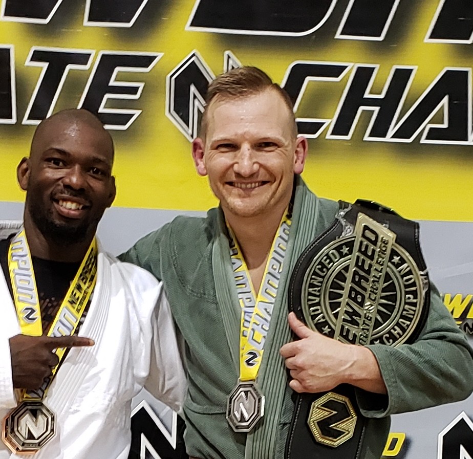 Two men posing for a photo, with one holding a medal and the other a large belt, in a martial arts setting.
